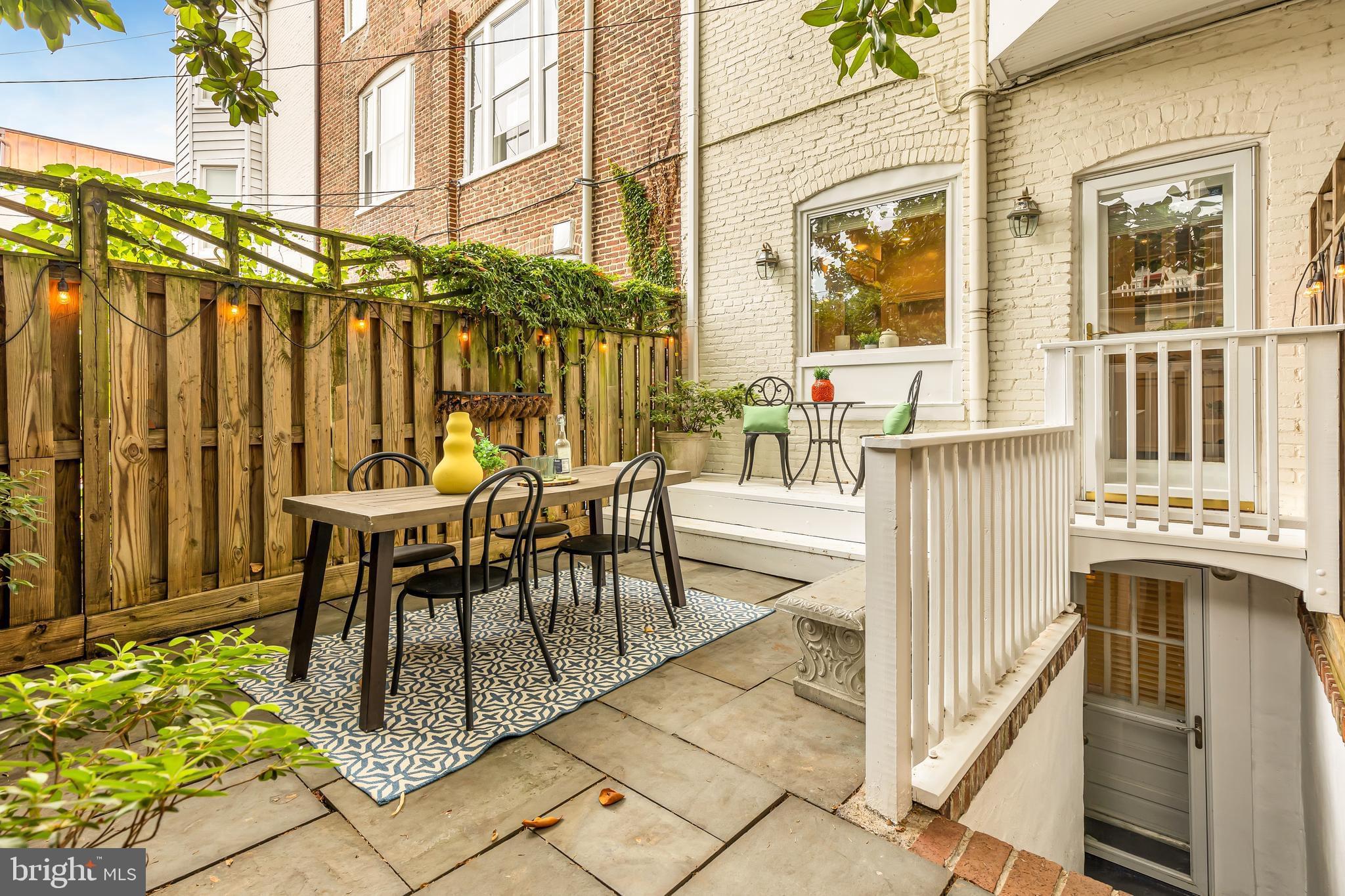 The back patio of a cream-colored brick townhouse with a wooden fence, outdoor furniture, and two access doors